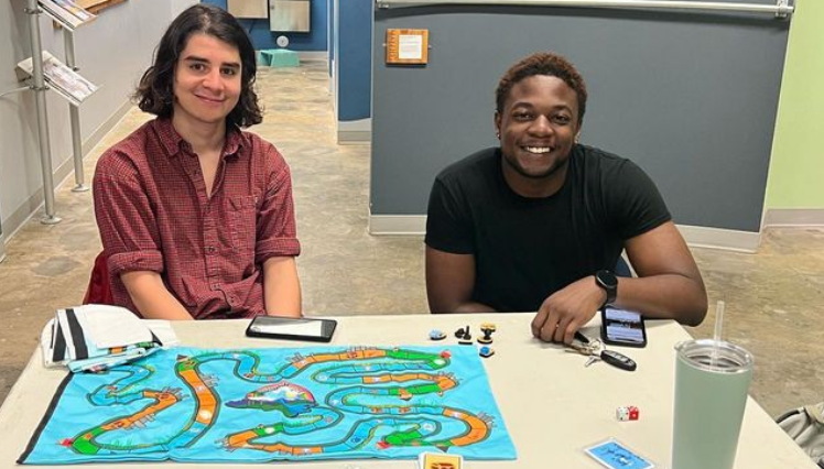 2 Climatopia students sitting behind a table and smiling with the Climatopia game board set up at the Kidzu Children's museum.