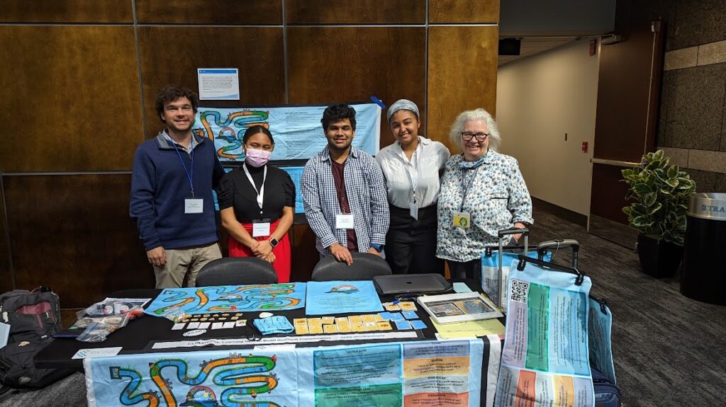 4 Climatopia students and Dr. Rachel Willis standing with their arms around one another behind their Climatopia set up at the CleanTech Summit.