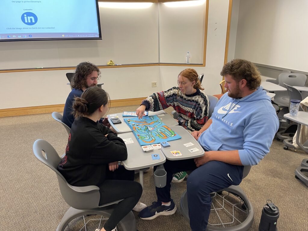 Rachel reaches out to move game piece while playing Climatopia with 3 other students