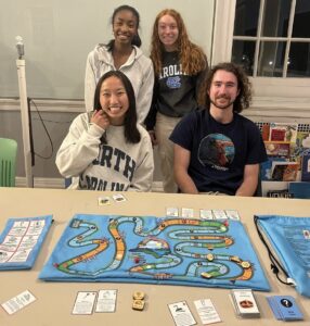 A group of students, Kailey, Rachel, Halle, and Jackson, pose with the Climatopia board game set on a table in front of them, at the Morehead Planetarium.