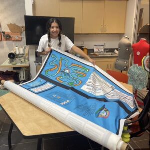 A young woman, Kim, holds a large roll of Climatopia fabric on a table in the BeAM Makerspace, preparing to cut and sew.