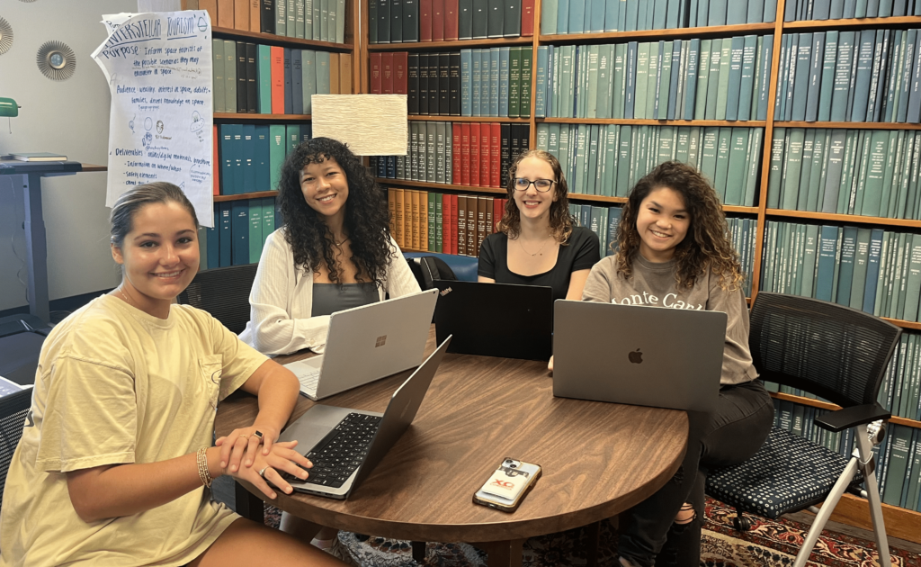 Four female college students are sitting in a library at a table with laptops.