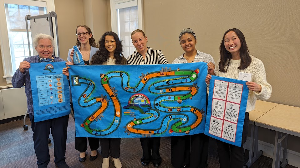 The Climatopia team (six women of various ages and races) stand holding up a game board.