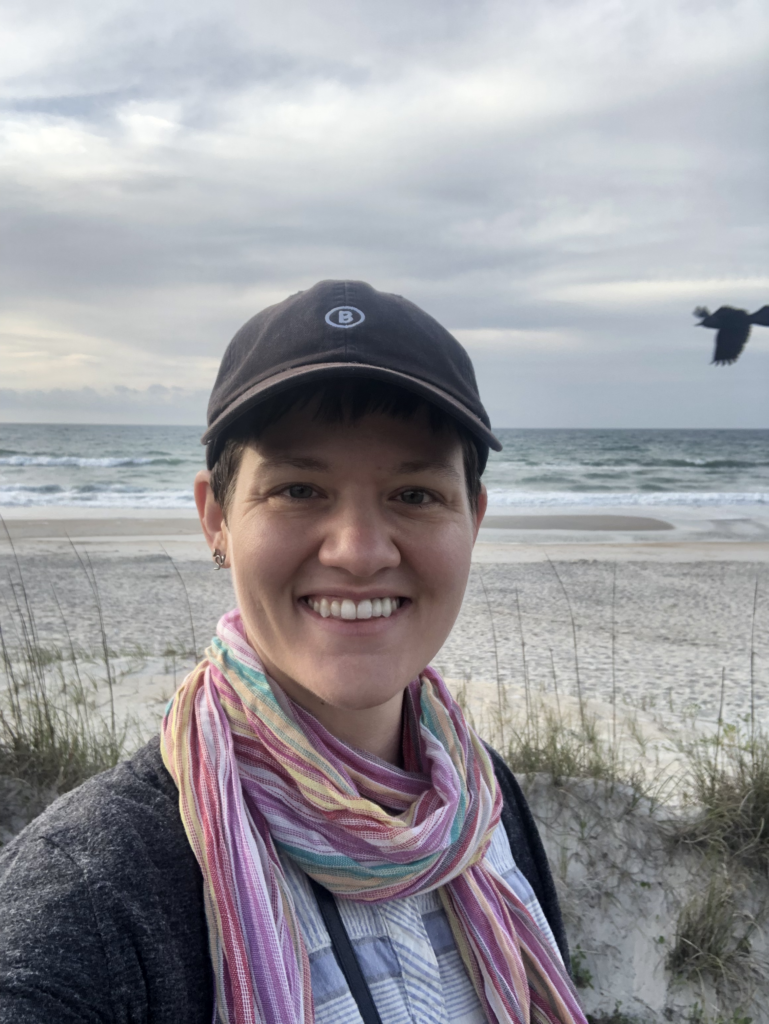 Jamie, a white woman, smiles at the camera at the coast. She is wearing a black baseball cap and a colorful scarf.