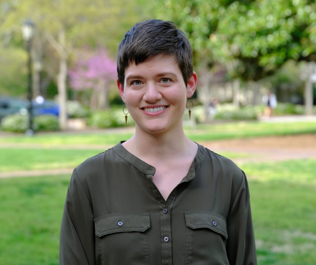 A white woman with short brown hair smiles at the camera as she stands in front of a campus path. She is wearing a green dress shirt and brass earrings.