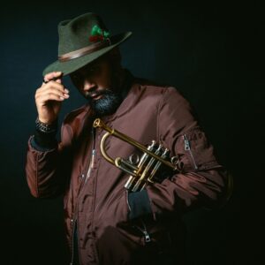 Wearing a green fedora and maroon bomber jacket, Al Strong poses in front of a darkly lit background while holding a trumpet.