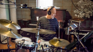 Dan Davis wearing studio headphones while adjusting his drums. Behind him three are two pianos and recording microphones.