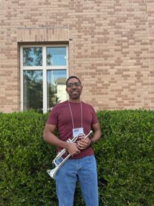 Samuel Edoho-Eket holds his trumpet outside of Kenan Music Building, in front of a hedge and a window. He wears a red shirt, jeans, and glasses.